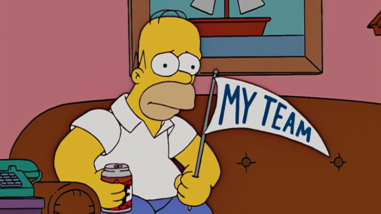 Homer Simpson sentado no sofá segurando uma cerveja e uma bandeira dos Simpsons