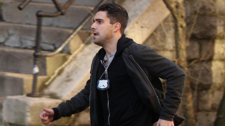 Fabien Frankel with slicked back brown hair running through the streets of New York. He's wearing a black hoodie underneath a gray jacket. There is a police officer's badge around his neck.