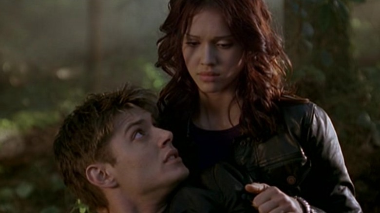 Jessica Alba wearing a black leather jacket with brown hair in Dark Angel, she's holding Jensen Ackles who is lying on the floor in a forest.