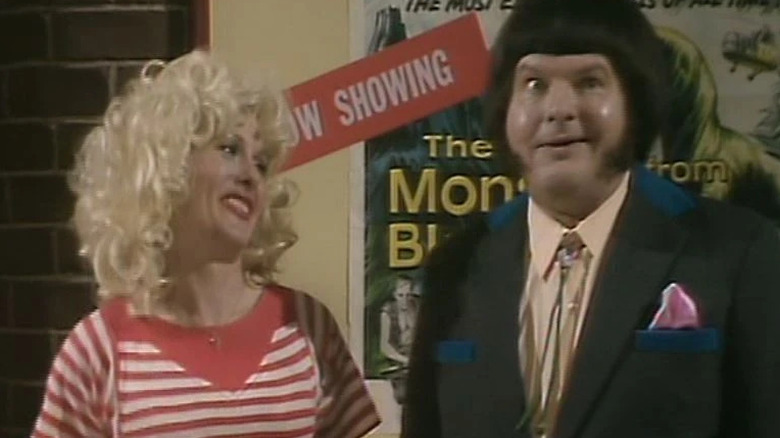 Ruthie (Jenny Lee Wright) stands smiling next to a mugging Harry (Benny Hill) in The Benny Hill Show