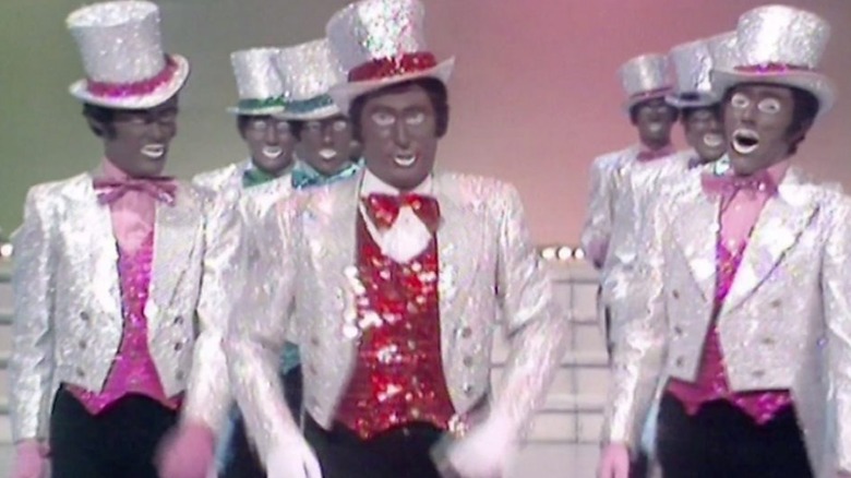 A group of performers in blackface wearing sequined suits and top hats sing in The Black and White Minstrel Show