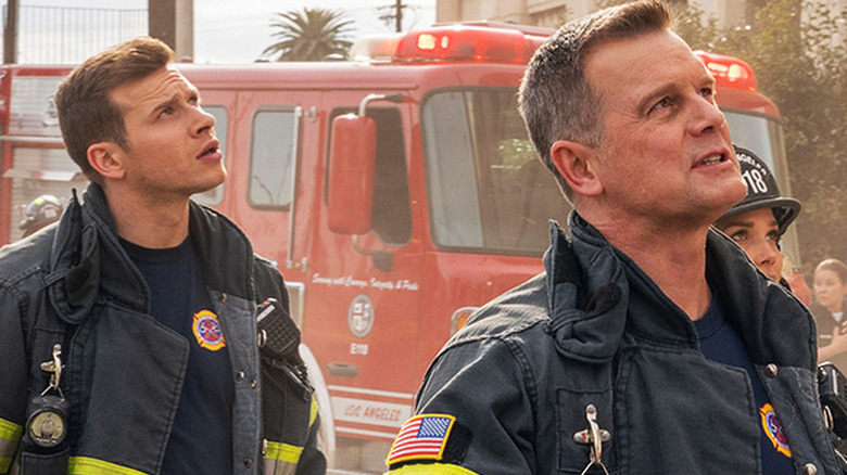 Oliver Stark as Buck and Peter Krause as Bobby in firefighter uniforms looking up in front of firetruck in 9-1-1