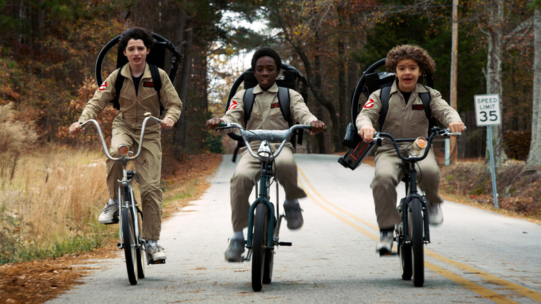 Mike, Lucas, and Dustin riding bikes in Ghostbusters costumes