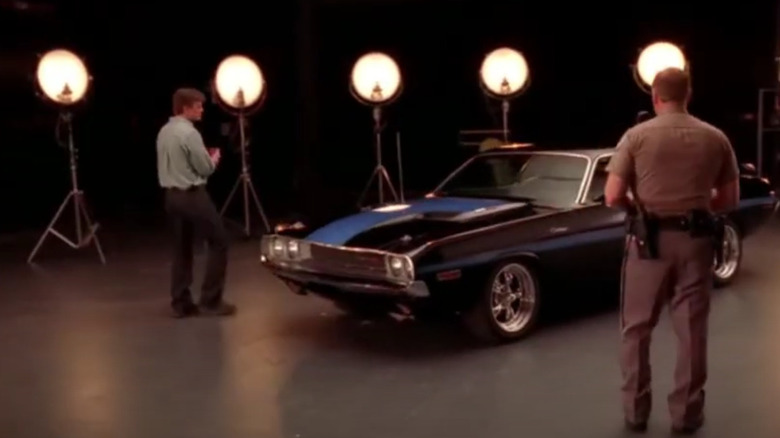 Nathan Fillion as Alex Tully circling a well-lit black car while a police officer observes on Drive