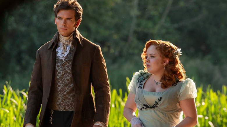 Luke Newton as Colin walking next to Nicola Coughlan as Penelope in Regency-era clothing in Bridgerton