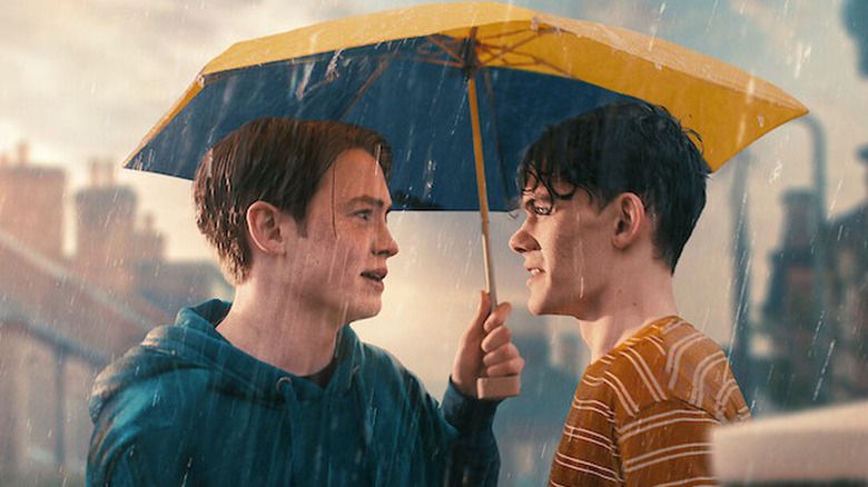 Kit Connor as Nick and Joe Locke as Charlie standing under umbrella in the rain in Heartstopper