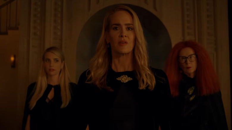 Three witches stand in the center of a large room