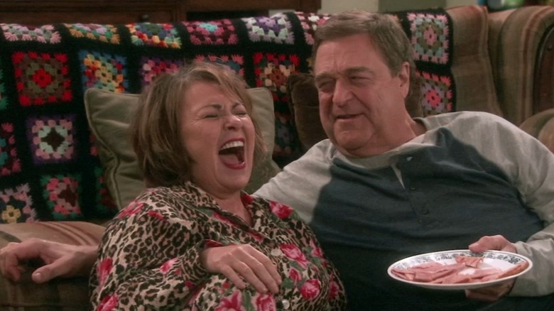 Roseanne as Roseanne Conner, and John Goodman as Dan Conner, laugh together on Roseanne, shortly before it rebooted as The Conners