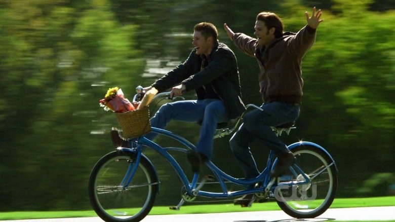 Sam Winchester (Jared Padalecki) outstretches his arms while riding on a tandem bicycle with Dean Winchester (Jensen Ackles) in Supernatural