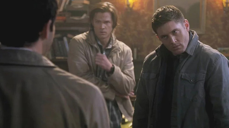 Dean Winchester (Jensen Ackles) hunches his shoulders while staring at Misha Collins as Sam Winchester (Jared Padalecki) watches in Supernatural
