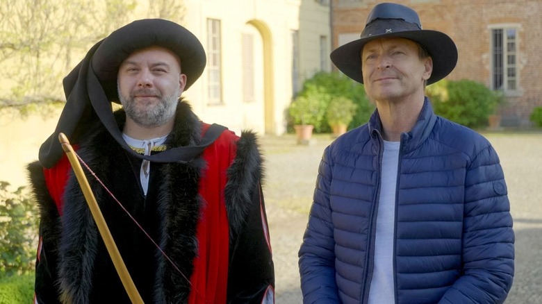 A man dressed in renaissance attire next to Phil Keoghan in "The Amazing Race" Season 38.