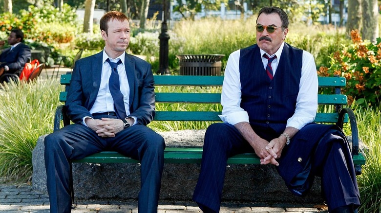 Donnie Wahlberg, dedektif Danny Reagan rolünde, mavi bir takım elbise ve beyaz yakalı bir gömlek giyiyor, Komiser Frank Reagan'ın yanında yeşil bir park bankında oturuyor, koyu lacivert bir yelek ve pantolon giyiyor, dizinde bir ceket ve mavi ve kırmızı çizgili bir kravat takıyor, Blue Bloods'da