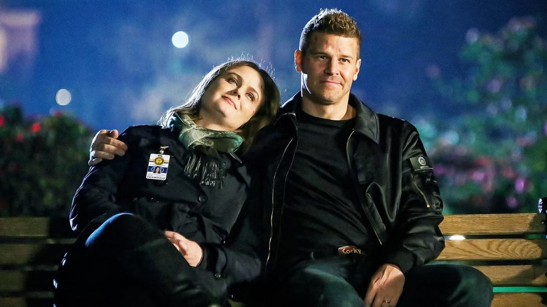 Emily Deschanel as Dr. Temperance "Bones" Brennan, wearing a black coat with a green scarf and her ID badge clipped to her coat, smiling and leaning her head on the shoulder of David Boreanaz as agent Seeley Booth, wearing a black jacket with a black shirt and jeans, who his arm around her and smiles as they sit on a park bench in Bones
