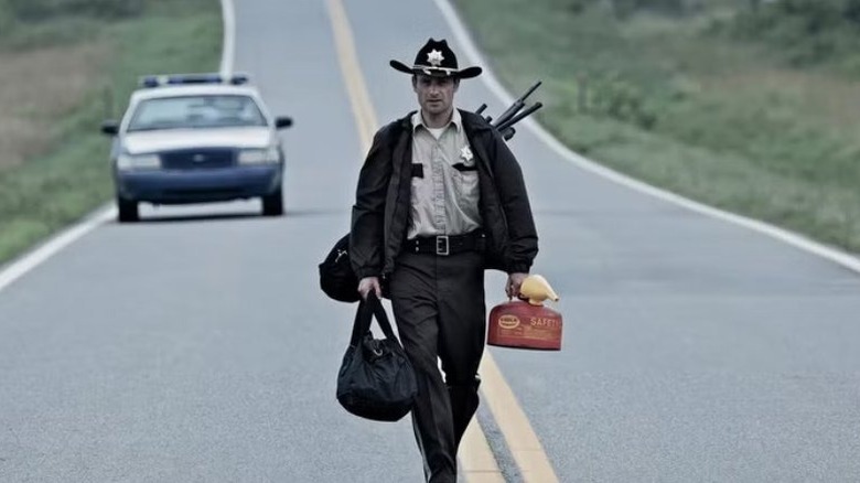 Rick Grimes elinde ve sırtında malzemelerle boş bir yolda yürüyor, polis arabasını arka planda bırakıyor