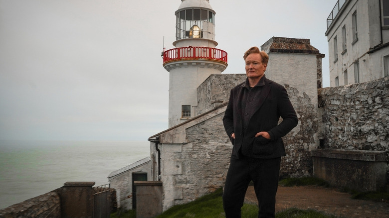 Conan O'Brien parado em frente a um farol com as mãos nos bolsos de seu terno de tweed cinza