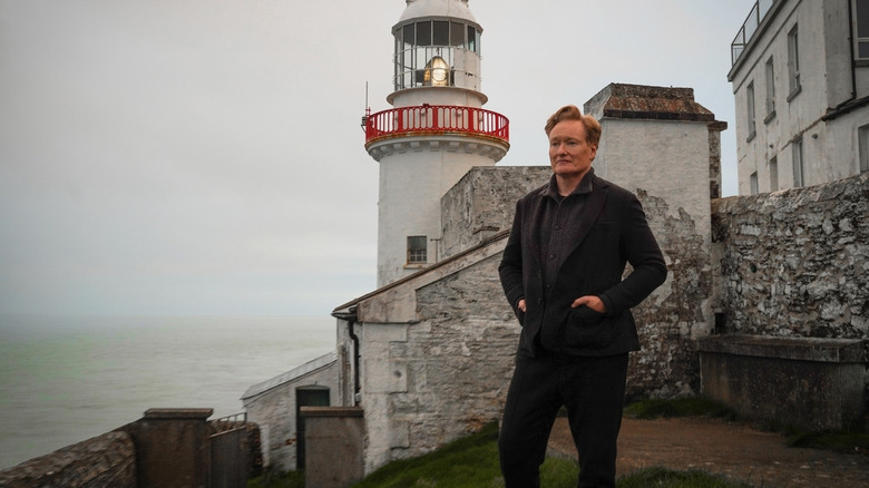 Conan O'Brien holds his hands in the pockets of his grey tweed suit while standing in front of a lighthouse in "Conan O'Brien Must Go"