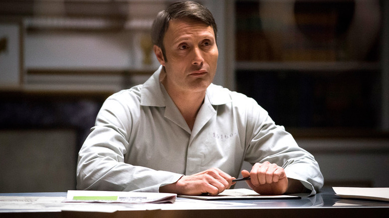 Hannibal Lecter (Mads Mikkelsen) sits at a table in a prison uniform in Hannibal