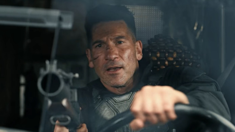 Jon Bernthal as the Punisher with black hair, driving a van and pointing a grenade launcher through a hole in the window.