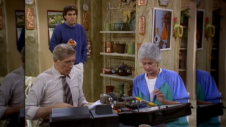 George Clooney eating and apple while Dorothy Zbornak and a man wearing a tie look a machinery