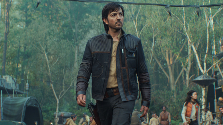 Cassian Andor caminha pelo acampamento rebelde em Yavine vestindo uma jaqueta de couro 