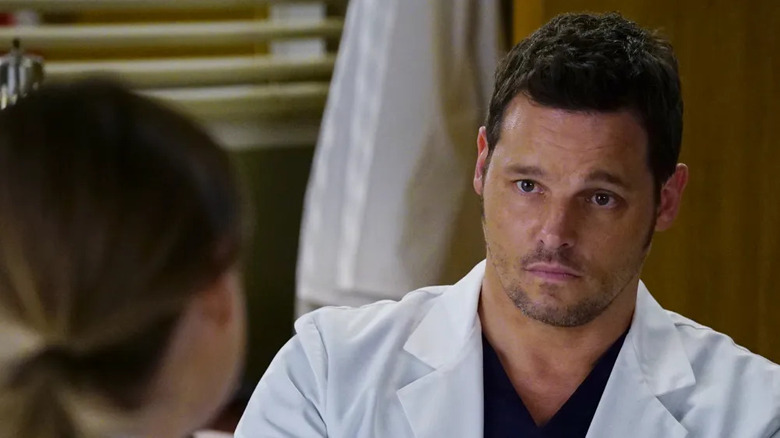 Justin Chambers, Grey's Anatomy'de sadece arkadan gördüğümüz, saçları at kuyruklu bir kadın karakterle konuşurken beyaz laboratuvar önlüğü ve siyah önlükle endişeli görünen Dr. Alex Karev rolünde