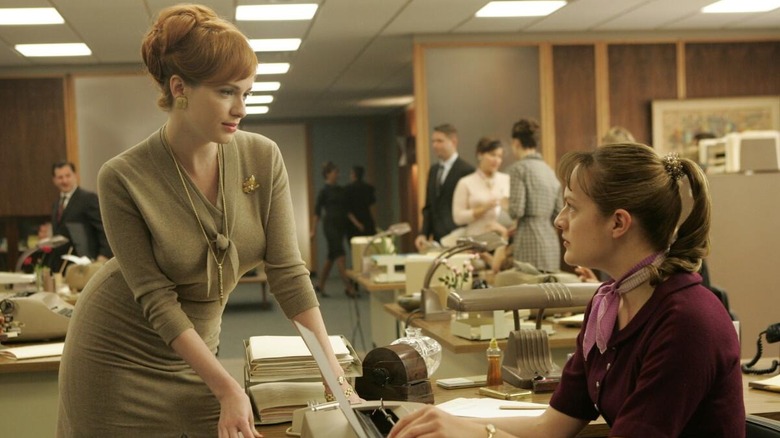 Joan (Christina Hendricks) stands leaning on a desk while Peggy (Elisabeth Moss) sits at a typewriter in the middle of a busy office setting in Mad Men