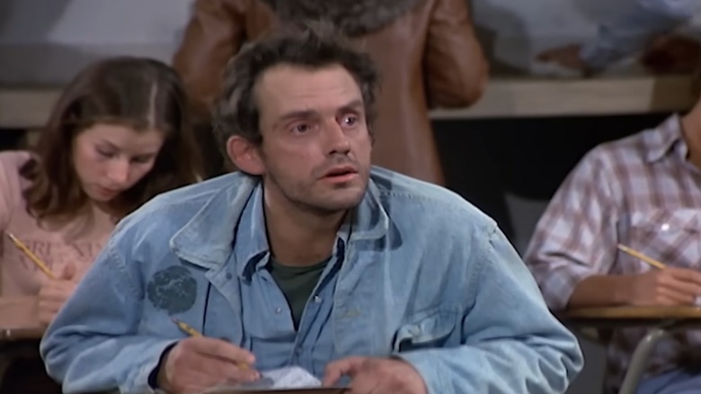 Iggy in "Taxi," wearing a denim jacket over a blue shirt as he writes on a school desk.