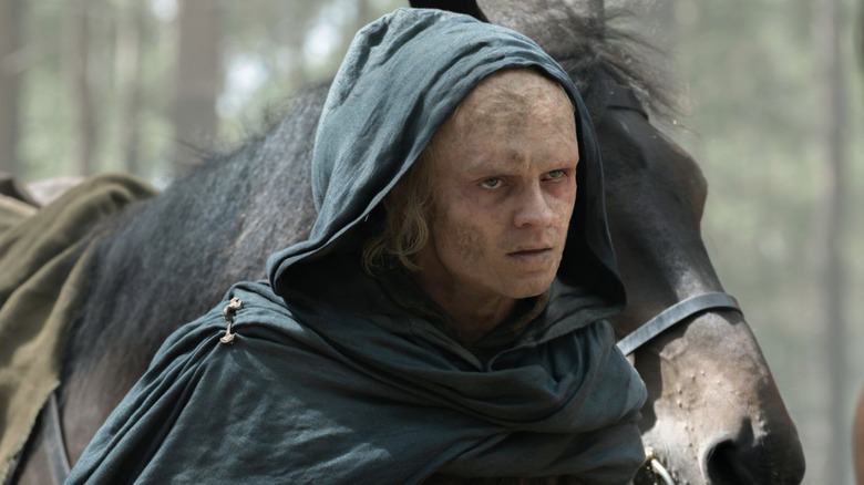 Tom Glynn-Carney as Aegon II Targaryen with a burnt face wearing a hood standing in front of a horse in House of the Dragon