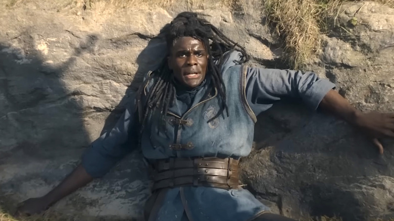 Clinton Liberty as Addam of Hull with black dreadlocks while wearing a blue tunic, he's lying against a large rock while the shadow of a dragon looms over him.