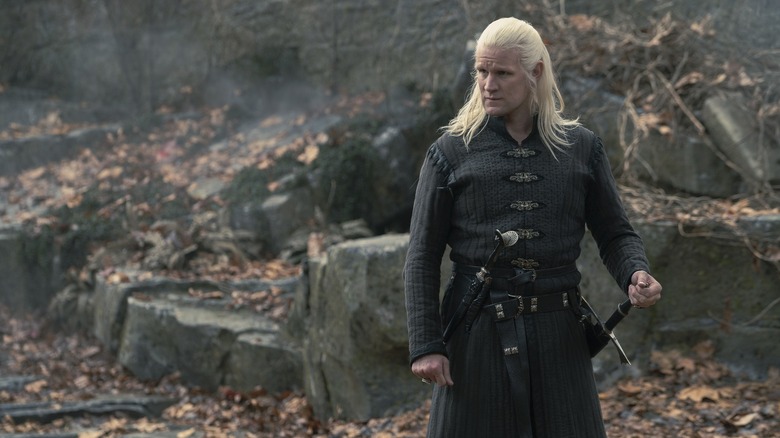 Matt Smith as Daemon Targaryen with long white hair, standing outdoors with rocks and fallen leaves behind him
