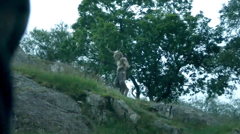An out-of-focus faun-like man with antlers and hooves stands on the top of a hill looking down.