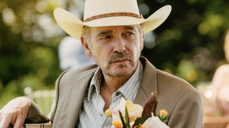 An older man with graying stubble wearing a gray suit, a gray and white striped shirt, and a beige cowboy hat with a brown rim. There are flowers in front of him.