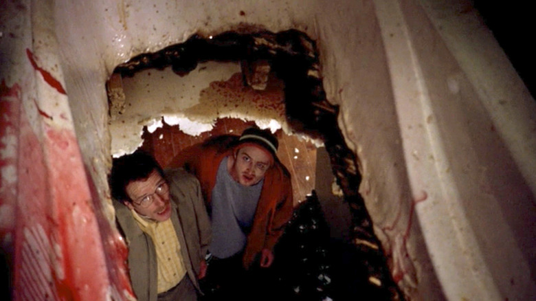 Jesse Pinkman, wearing a beanie and red sweater, and Walter White, wearing a yellow shirt and jacket, look up at the hole in their ceiling leading to bloody bathtub in "Breaking Bad"
