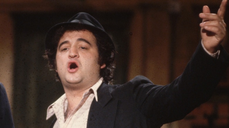 John Belushi wearing a black fedora wearing a black suit and white dress shirt without a tie.