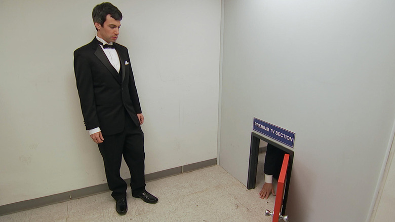 Nathan Fielder usa smoking enquanto fica ao lado de um homem ajoelhado do outro lado de uma pequena porta chamada Seção de TV Premium. 