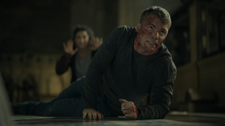 Peter Sutherland blood-soaked and crawling on his elbows while Freya holds her hands up in the background in The Night Agent