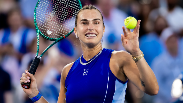 Aryna Sabalenka in a blue tennis shirt holding a racket and tennis ball