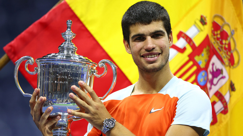 Carlos Alcaraz holding the U.S. Open Championship trophy in front of a Spanish flag