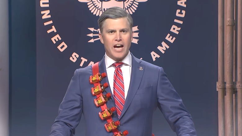 Colin Jost poses as Pete Hegseth wearing a blue blazer, red and blue tie and a sash containing fake alcohol on "Saturday Night Live."