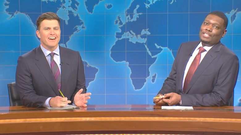 Colin Jost and Michael Che wear ordinary suits and sit behind a brown desk on "Saturday Night Live."