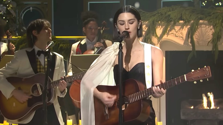 Gracie Abrams playing a guitar in a black dress in front of a band