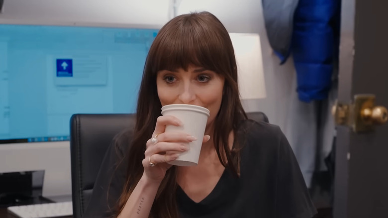 Dakota Johnson drinks from a white cup in an office in "Saturday Night Live"