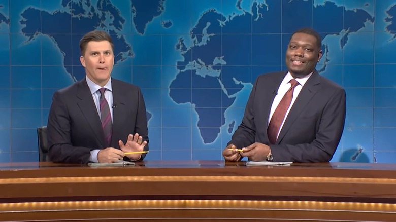 Colin Jost and Michael Che sit in suits behind a desk and in front of a world map for "Saturday Night Live."