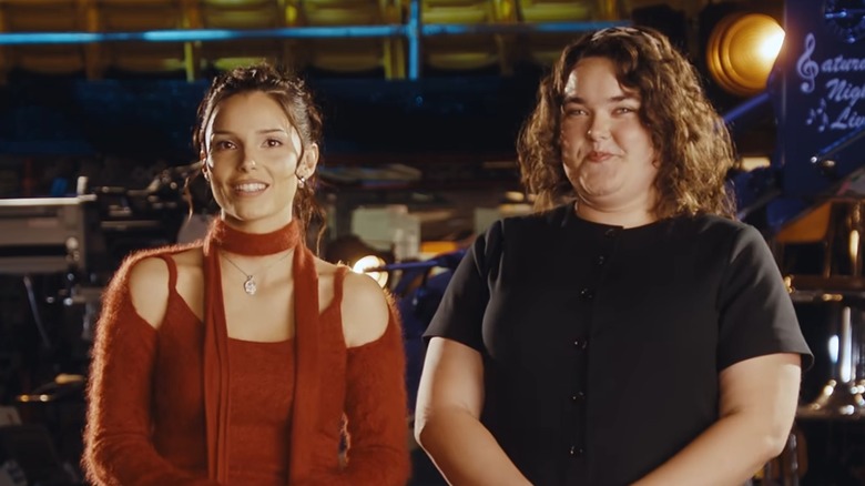 Inde Navarrette in a red collarless and shoulderless sweater next to Grace Reiter in a black shirt on the "Saturday Night Live" stage.