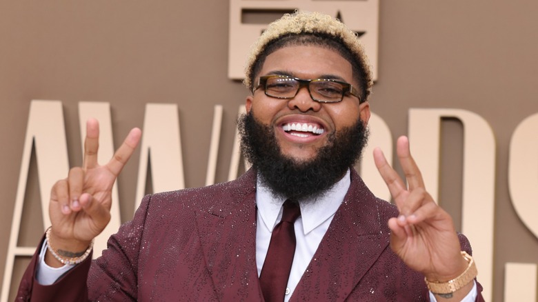 Druski holding up peace signs while wearing a burgundy suit at the BET awards.