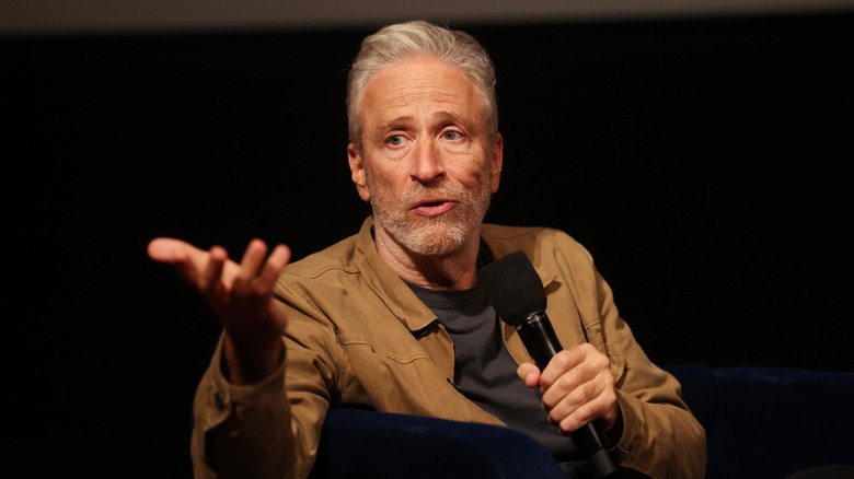 Jon Stewart with a microphone, seated and talking on stage