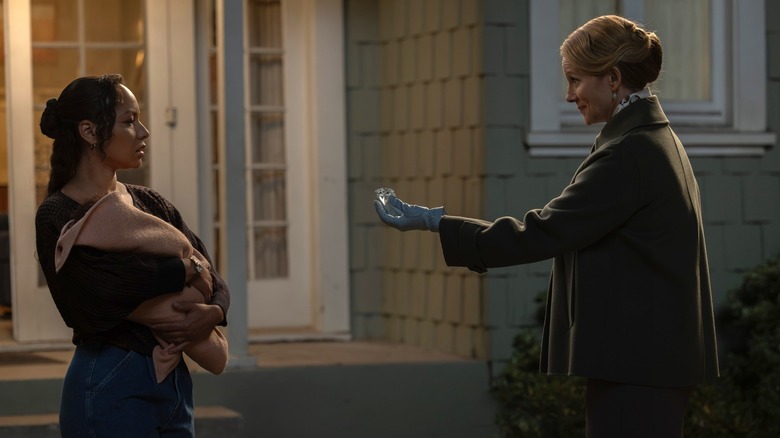 Two women. On the left is Jasmine Cephas Jones as Bernadette Stewart. She's stood outside of her house, she has long brown curly hair which is tied back. She's wearing a black and brown sweater with blue jeans and she's holding a baby. On the right is Laura Linney as the human form of the Guardian. She has tied back blond hair and is wearing a long green coat. She's also wearing a light blue leather glove on her left hand, which is holding out a diamond towards Bernadette.