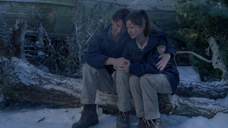 Detective Woody Hoyt hugging Dr. Jordan Cavanaugh as they sit on a snowy tree trunk in the woods