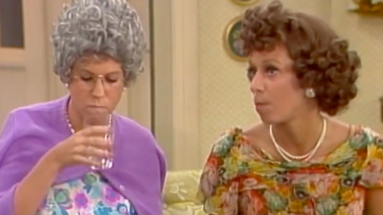 Mama (Vicki Lawrence) sips from a glass with a stern, pursed expression beside Eunice (Carol Burnett), who looks off to the side with her mouth slightly open as if reacting to someone offscreen in Mama's Family