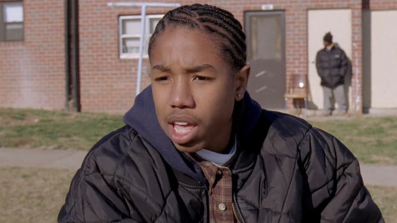 Michael B. Jordan wearing a quilted black jacket with a blue hood and a plaid shirt underneath as Wallace in "The Wire."